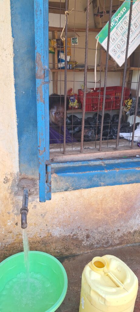Caption: Zura Akinyi sits inside the newly restored community water kiosk, operating the kiosk and serving customers with safe, reliable water. Photo credit: Susan Kanga.